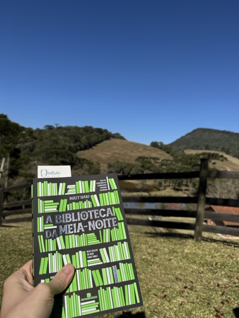 Eu segurando o livro com uma paisagem de fundo. Baseado nessa leitura fiz a resenha de A Biblioteca da Meia-noite.